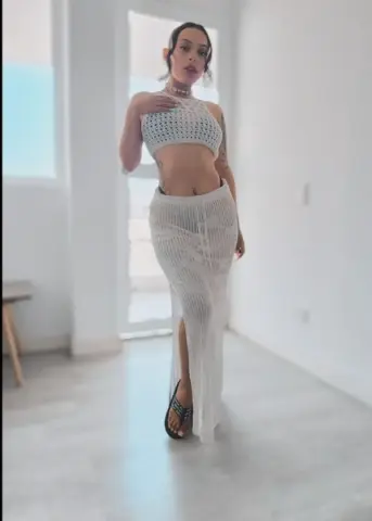 Young woman in a white crochet crop top and matching maxi skirt standing in a bright, minimalist room.