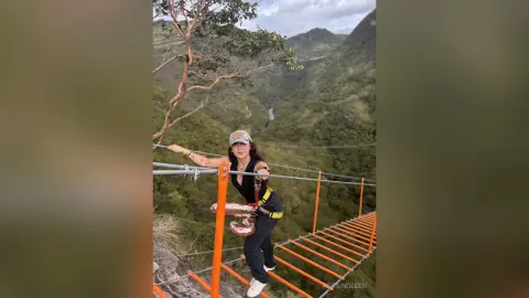 RossiNoleen stands on a narrow orange suspension bridge overlooking a lush, green mountain valley.