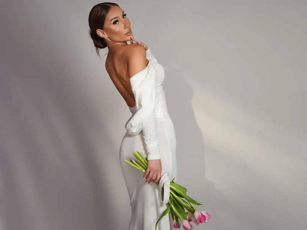 Elegant woman in a white long-sleeve gown holding pink tulips against a minimalist studio background.