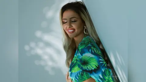 Anita smiles with her eyes closed, wearing a green tropical leaf print top and large hoop earrings against a white wall.