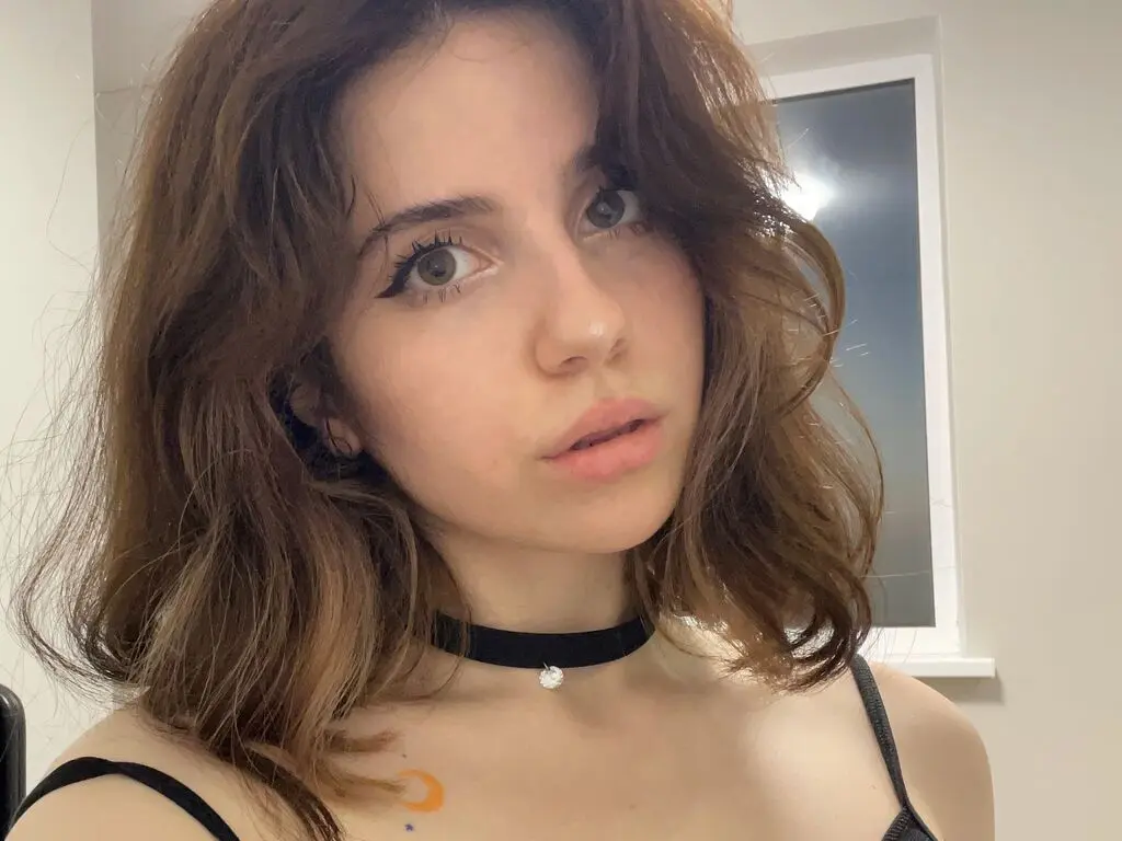 Young woman with wavy brown hair wearing a black choker and tank top in a bright indoor setting.