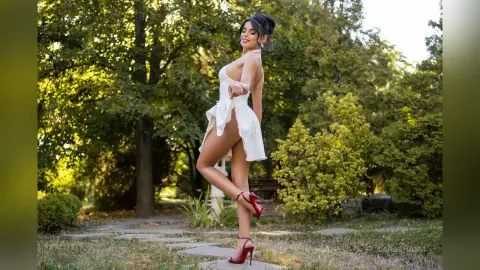 Smiling woman in a white mini dress and red heels posing in a sunny green garden setting.