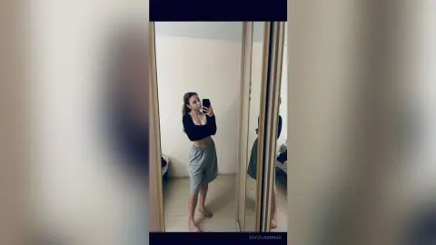 Young woman in a black crop top and grey sweatpants taking a mirror selfie in a bright room.