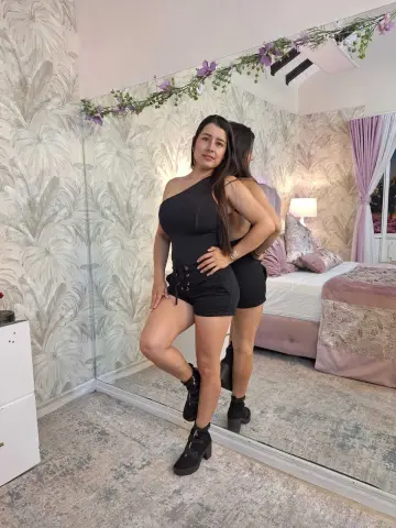 Woman in black romper and boots posing in a bright bedroom with floral wallpaper and a large mirror.