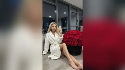Blonde woman in a white bathrobe sitting near a large bouquet of red roses in a modern living room.