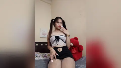 Young woman in a white lace corset and black skirt posing in a cozy bedroom with a red teddy bear.