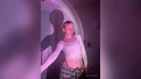Young woman in a white long-sleeve crop top and plaid skirt posing under dreamy purple rainbow lighting.