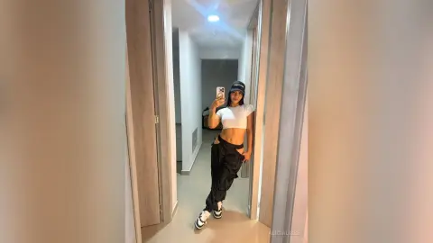 Young woman in white crop top and black cargo pants taking a mirror selfie in a modern hallway.