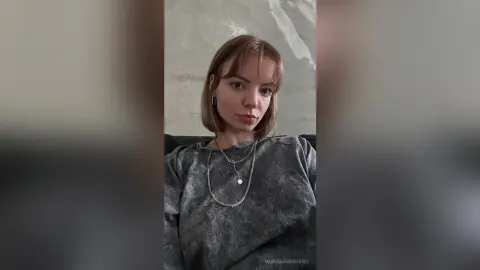 Young woman with short blonde hair wearing a grey tie-dye sweatshirt and silver necklaces in a minimalist room.