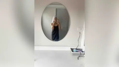 Young woman in a black towel and white hair wrap taking a selfie in a bright, minimalist bathroom mirror.
