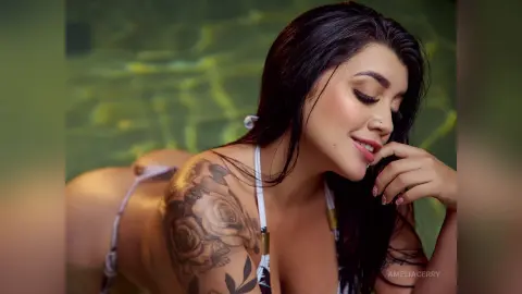 Smiling woman with floral tattoos in a white bikini relaxing by a green swimming pool.