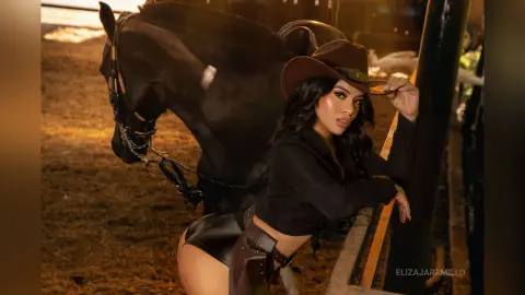 Beautiful woman in black outfit and cowboy hat posing with a dark horse in a rustic stable setting.