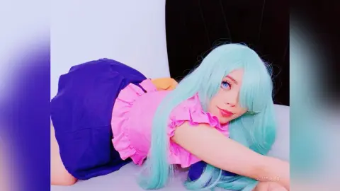 HimeJess poses on her stomach wearing a pink ruffled top and blue shorts, showing off her long light blue hair and blue eyes.