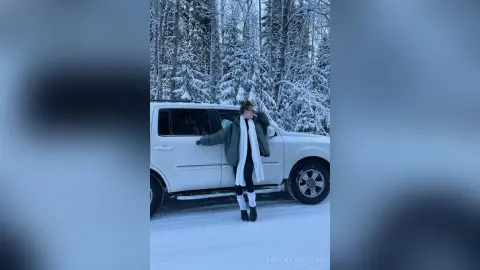Woman in a white scarf leaning on a white SUV in a snowy winter forest setting.