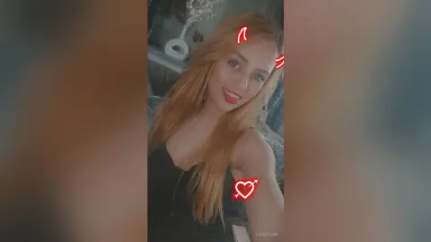 Smiling blonde woman in a black tank top posing for a casual selfie in a cozy indoor setting.