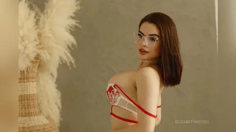 Young woman with glasses in red and white lace lingerie posing in a minimalist neutral room.