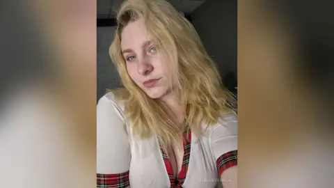 StellaVanilla poses with her long blonde hair down, wearing a white top with red plaid trim.
