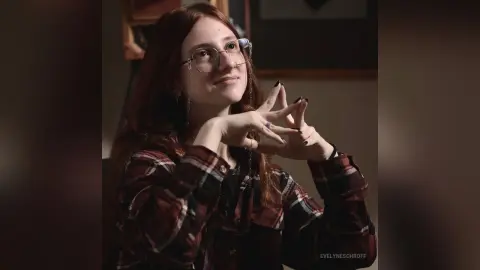 Smiling young woman with glasses and red hair wearing a plaid flannel shirt in a cozy indoor setting.