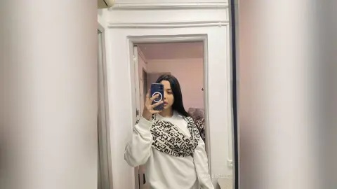 Young woman in a white Fendi sweatshirt taking a mirror selfie in a bright minimalist room.