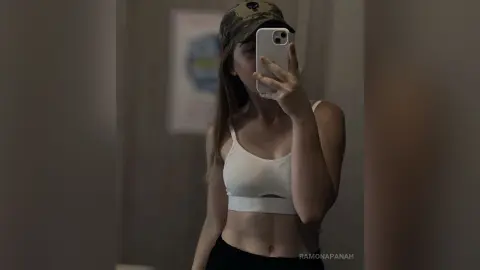 Young woman in a white sports bra and camo hat taking a moody mirror selfie in a dimly lit room.