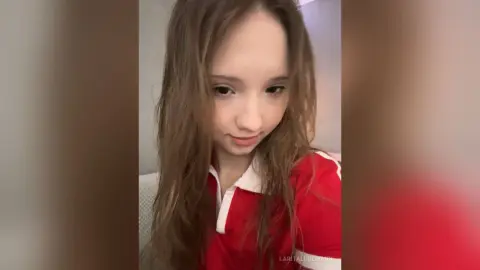 LaritaLudemann looks down with long brown hair while wearing a red and white collared shirt.