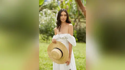 LucyMartin poses outdoors in a white off-the-shoulder top while holding a large straw hat.