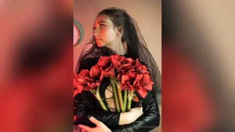 Moody portrait of a woman in a black outfit holding a large bouquet of red flowers with a sheer black veil.