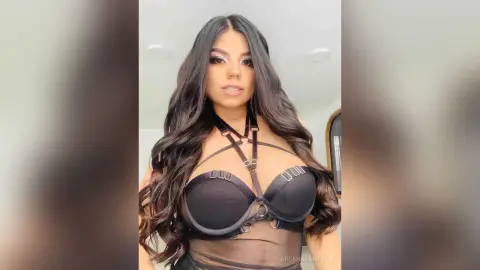Glamorous woman with long wavy hair wearing a black strappy bodysuit in a bright indoor setting.
