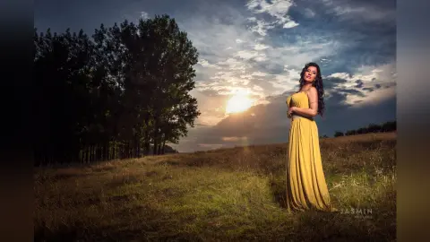 MissyJolie stands in a grassy field wearing a long, strapless yellow dress against a sunset sky.