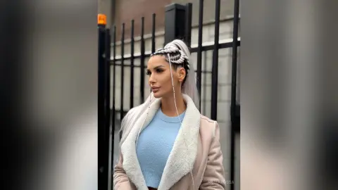 Stylish woman in a light blue sweater and beige shearling coat posing outdoors near a black metal fence.
