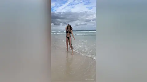 Woman in a black cutout swimsuit walking on a cloudy beach under a dramatic sky.