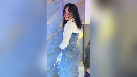 NaomiVelour poses sideways, showing her curves in a white long-sleeve shirt and a high-waisted denim skirt with her long, dark curly hair.