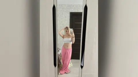 Blonde woman in white crop top and pink wide-leg trousers posing in a modern minimalist bedroom mirror.
