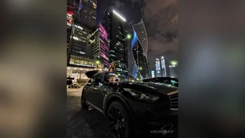 Stylish woman in casual attire standing by a black luxury car in a glowing urban cityscape at night.