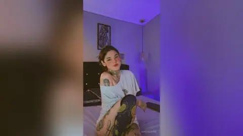 Young woman with tattoos in a white t-shirt sitting in a moody, purple-lit bedroom.