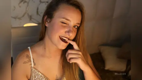 Smiling young woman in a sparkly sequin top posing playfully in a cozy, dimly lit bedroom setting.