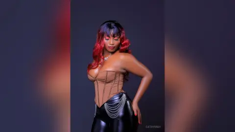 Glamorous woman with red hair in a brown corset and black leather pants posing against a moody studio background.