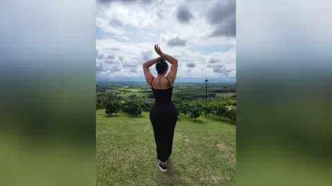 Curvy woman in a black dress standing in a lush green field under a cloudy sky, enjoying a peaceful landscape.