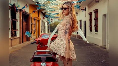 Stylish woman in a floral sundress and sunglasses posing with a red scooter on a sunny, decorated cobblestone street.