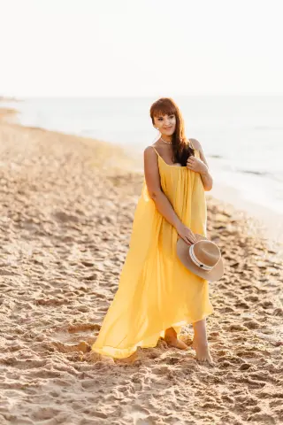 Woman in a flowing yellow maxi dress holding a straw hat on a sunny beach at sunset.