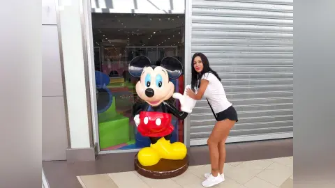 Smiling woman in a white t-shirt and black shorts posing next to a large Mickey Mouse statue at a shopping mall.