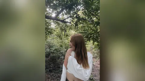 Young woman in a white shirt looking away under lush green forest trees in a peaceful outdoor setting.