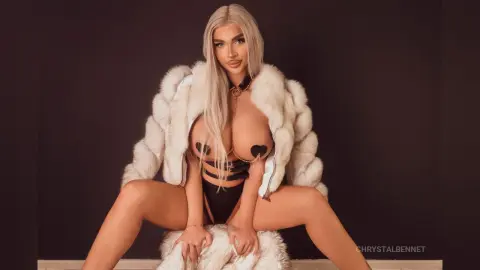 Blonde woman in black lingerie and white faux fur coat sitting against a dark background with a glamorous mood.