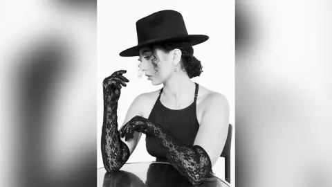 Elegant woman in black wide-brimmed hat and lace gloves posing in a minimalist black and white studio setting.