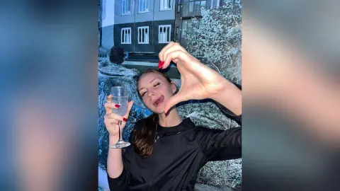 Cheerful young woman in a black jacket holding a glass of bubbly outdoors at night near snowy bushes.