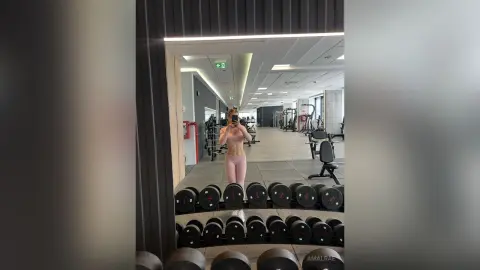 Fit woman in pink gym wear taking a mirror selfie in a modern bright fitness center with dumbbells.