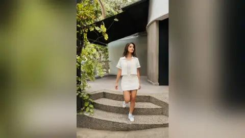 TefyRusso stands on concrete steps wearing a white short-sleeved crop top and a matching white mini skirt, looking off to the side with long brown hair.