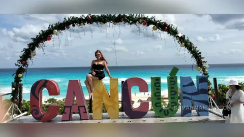 Stylish woman in a black dress sitting by large Cancun sign on a sunny tropical beach with ocean view.