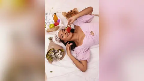 Smiling woman in a pink summer dress enjoying a picnic with fruit and flowers on a white blanket.