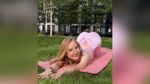 Smiling blonde woman in pink yoga outfit relaxing on a pink mat in a sunny green grass field.